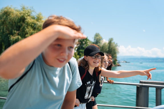 Gruppe von Jugendlichen steht an einem Gel&auml;nder am Seeufer, blickt aufs Wasser und zeigt auf etwas in der Ferne an einem sonnigen Tag.