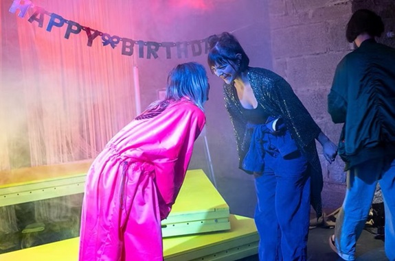 Farbig beleuchtete Szene mit mehreren Personen; im Vordergrund stehen sich zwei Personen gegen&uuml;ber, im Hintergrund h&auml;ngt ein &bdquo;HAPPY BIRTHDAY&ldquo;-Banner.