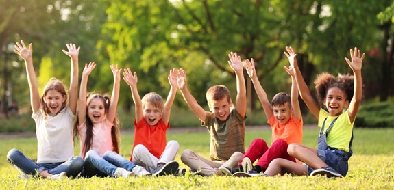 Mehrere Kinder sitzen im Freien auf einer Wiese und heben beide Arme in die Luft.