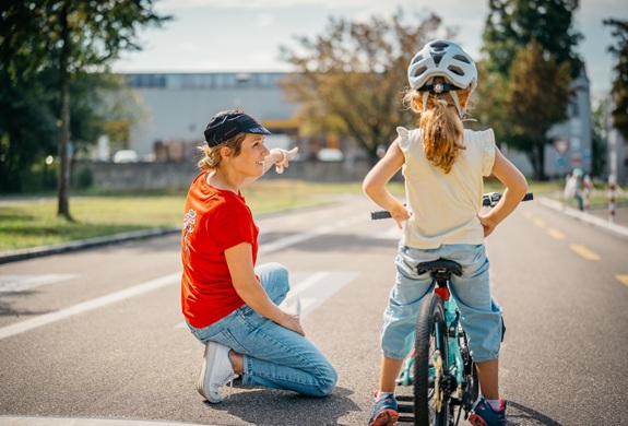 Person kniet auf einer Strasse und spricht mit einem Kind auf einem Fahrrad w&auml;hrend eines &Uuml;bungskurses im Freien.
