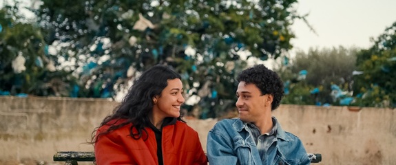Filmszene: Zwei Personen sitzen nebeneinander auf einer Bank vor einer Mauer mit B&auml;umen im Hintergrund.