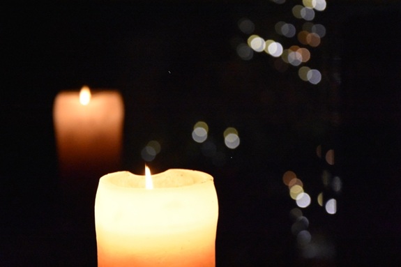 Zwei brennende Kerzen vor dunklem Hintergrund mit unscharfen Lichtpunkten im Hintergrund.