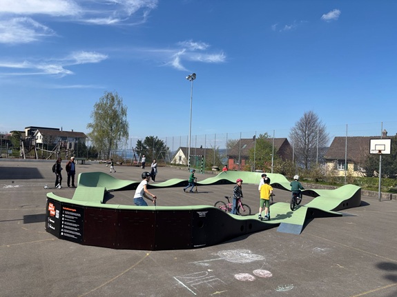 Ein Pumptrack auf einem asphaltierten Platz, auf dem mehrere Kinder mit Fahrr&auml;dern und Scootern fahren. Im Hintergrund sind H&auml;user, ein Basketballkorb und ein Zaun zu sehen,.