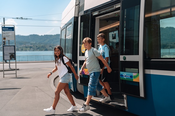 Drei Kinder steigen aus einer blauen Stra&szlig;enbahn an einer Haltestelle aus. Im Hintergrund sind ein See, Berge und ein Fahrplan an der Haltestelle zu sehen.