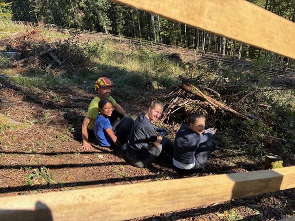 Eine Gruppe sitzt im Wald auf dem Boden, begleitet von einer Person mit Schutzhelm.