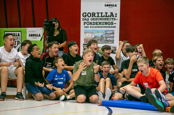 Eine Gruppe Kinder sitzt jubelnd in einer Turnhalle und reagiert begeistert auf ein Ereignis.