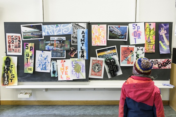 Kind betrachtet eine Ausstellung mit bunten Plakaten, Fotos und Graffiti-Arbeiten an einer Pinnwand. Die kreativen Werke zeigen verschiedene Schriftz&uuml;ge und Kunstprojekte.