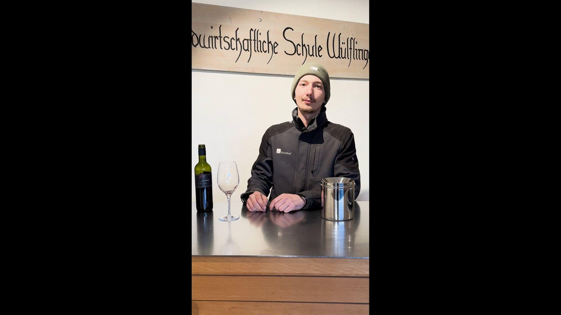 Ein Mann mit Kappe und dunkler Jacke steht hinter einem Tresen. Auf dem Tresen steht eine Flasche Wein, ein Weinglas sowie ein  Crachoir.