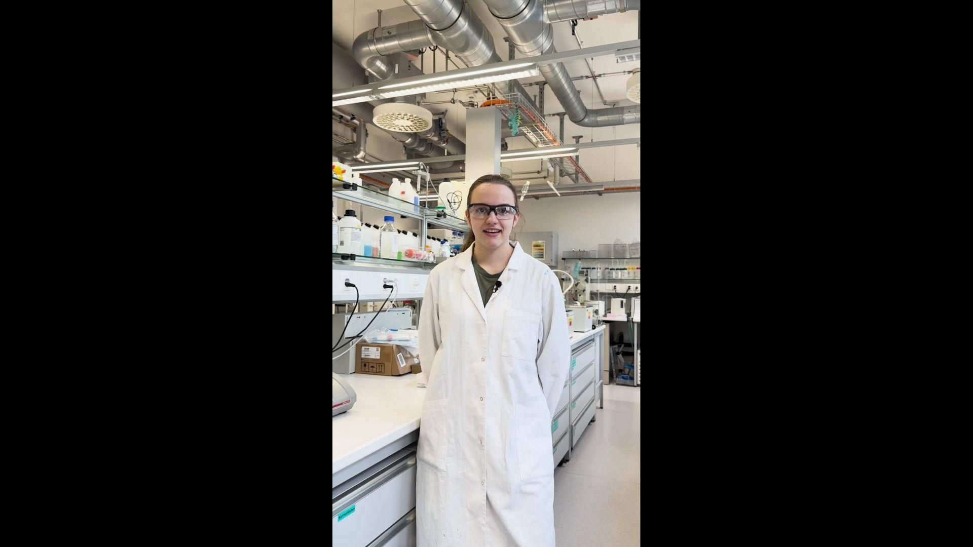 Eine Frau mit langen, zurückgebundenen dunklen Haaren steht in einem Labor. Sie trägt einen weissen Laborkittel und eine Sicherheitsbrille. Im Hintergrund sieht man eine lange Arbeitsbank mit zahlreichen kleinen Plastikflaschen.