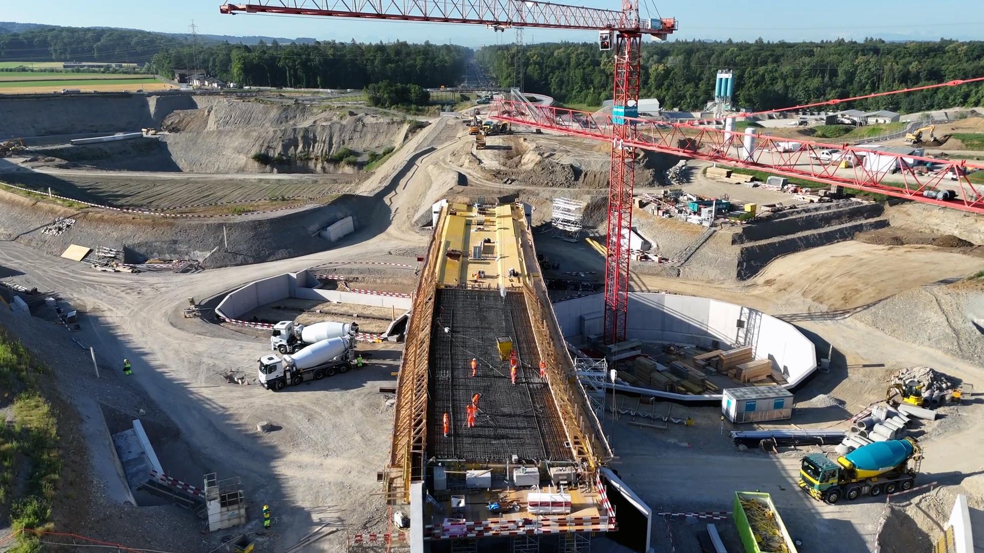 Ein Drohnenfoto zeigt eine große Straßenbaustelle. Im Vordergrund wird eine Autobahnbrücke gebaut, auf der Bauarbeiter zu sehen sind. Rechts steht ein roter Kran, und links befindet sich eine ausgehobene Grube. Im Hintergrund erstreckt sich eine neue Straße durch ein Waldgebiet. Am unteren Bildrand stehen Betonmischer und weitere Baufahrzeuge.